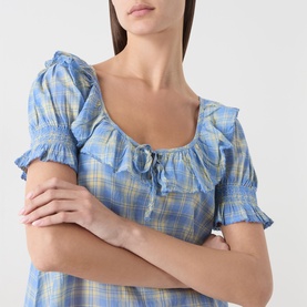 Model wearing Damson Madder Harlow Check Mini Dress in blue check, showing ruched square neckline, self-tie detail and shirred puff sleeves
