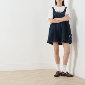 Model wearing Damson Madder Immy Denim Mini Dress in blue with pleated details and adjustable straps, styled with white tee and loafers