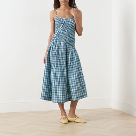 Model wearing Damson Madder Lyra Panelled Tomboy Check Dress in blue check, midi length with shirred bodice and flared skirt, styled with yellow mules