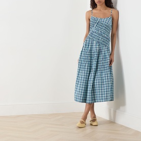 Model wearing Damson Madder Lyra Panelled Tomboy Check Dress in blue check, midi length with shirred bodice and flared skirt, styled with yellow mules