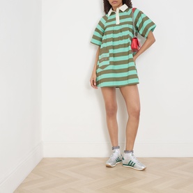 Model wearing Damson Madder Maggie Polo Dress in green multi stripe with white polo collar, styled with green trainers and red mini bag.
