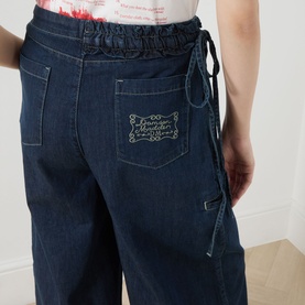 Model wearing Damson Madder Augusta Mid-Rise Straight Leg Jeans in dark indigo with frill tie waist and logo patch pocket.