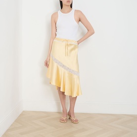 Model wearing Damson Madder Delphia Embellished Yellow Skirt with lace trim and asymmetric hem, styled with white vest and gold sandals.