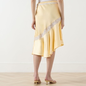 Model wearing Damson Madder Delphia Embellished Skirt in yellow satin with lace trim and asymmetric hem, styled with yellow heeled mules.