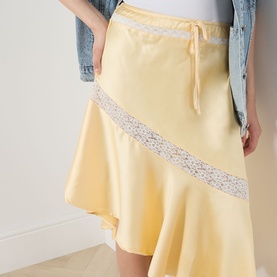 Model wearing Damson Madder Delphia Embellished Skirt in yellow satin with lace trim detail and asymmetric hem, styled with denim jacket