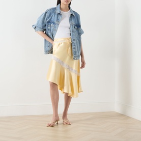 Model wearing Damson Madder Delphia Embellished Yellow Skirt with asymmetric hem and lace trim, styled with denim jacket and tan sandals.