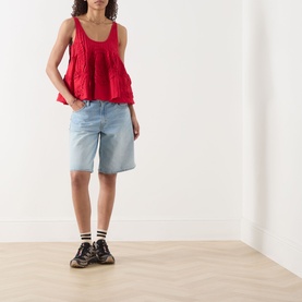 Model wearing Damson Madder Stella Ruffle Cami in red with scoop neck and flared hem, styled with light blue denim shorts and trainers.