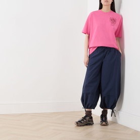 Model wearing Damson Madder Frida Bloomer Trousers in navy, with elasticated waist and self-tie cuffs, styled with a pink tee and sandals.
