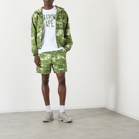 Model wearing BAPE Pixelated Camo Print Logo Windbreaker in green, styled with matching camo shorts, white tee, and grey trainers.