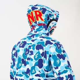Model wearing blue camouflage zip-up hoodie with branded hood graphic in red and yellow lettering, back view