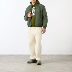 Model wearing olive quilted bomber jacket over green graphic hoodie, styled with cream wide-leg trousers and khaki trainers