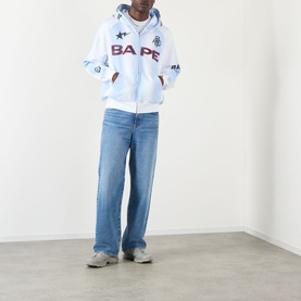 Model wearing BAPE Hand Drawn Football Zip Hoodie in white with star motifs, styled with light blue wide-leg jeans and grey trainers.