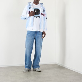 Model wearing BAPE Hand Drawn Football Zip Hoodie in white, styled with a graphic tee, light blue wide-leg jeans and grey trainers.