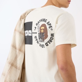 Model wearing BAPE A Bathing Ape Logo T-Shirt in ivory, showing back graphic print, styled with a plaid shirt draped over shoulder.