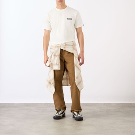 Model wearing BAPE A Bathing Ape Logo T-Shirt in ivory, styled with tan plaid shirt tied at waist, brown cargo trousers and chunky boots.