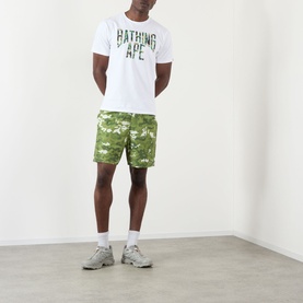 Model wearing BAPE Camouflage NYC Logo T-Shirt in white with green camo print logo, styled with matching green camo shorts and grey trainers.
