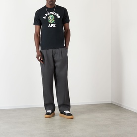 Model wearing BAPE ABC Camouflage College Logo Shirt in black with green camo motif, styled with grey trousers and two-tone trainers.