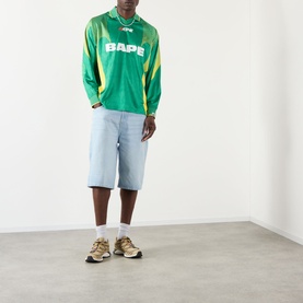 Model wearing BAPE Logo Football Jersey Long-Sleeved T-Shirt in green with yellow panels, styled with light blue denim shorts and gold trainers.