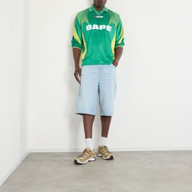 Model wearing BAPE Logo Football Jersey Long-Sleeved T-Shirt in green with yellow panels, styled with light blue denim shorts and tan trainers.