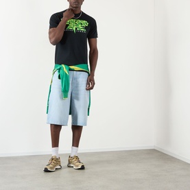 Model wearing BAPE Groovy Justice Logo T-Shirt in black with neon green graphic print, styled with light wash denim shorts and gold trainers.