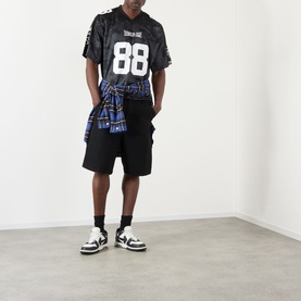 Model wearing BAPE Logo Football Jersey T-Shirt in black, number 88, styled with black shorts, plaid shirt tied at waist, and white trainers.