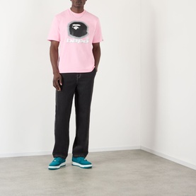 Model wearing BAPE Spray Ape Head Logo T-Shirt in pink, styled with black wide-leg trousers and teal trainers.