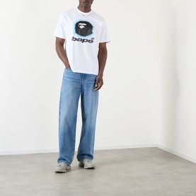 Model wearing BAPE Spray Ape Head Logo T-Shirt in white with blue ape graphic, styled with light wash wide-leg jeans and grey trainers.
