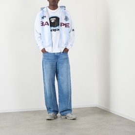 Model wearing BAPE Spray Ape Head Logo T-Shirt in white, styled with a light blue zip hoodie, wide-leg blue jeans and grey trainers.
