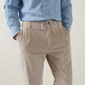 Model wearing Eleventy sand cotton trousers with drawstring waist and pleated front, styled with a blue linen shirt.