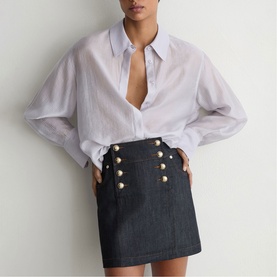 Model wearing Reiss dark blue denim mini skirt with gold button details, styled with a white open-collar oversized shirt