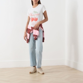 Model wearing Free People Cherry Combo graphic tee with light blue jeans, plaid shirt tied at waist, and cream trainers.