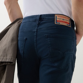 Model wearing Diesel 3049 tapered jeans in blue, close-up of rear view showing branded patch detail, styled with white tee and grey jacket