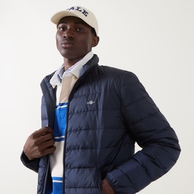 Model wearing Gant Evening Blue quilted down casual jacket with logo badge, styled with blue stripe polo and cap.