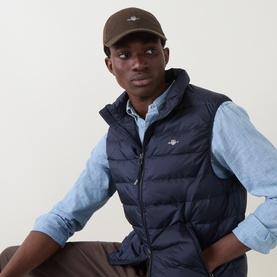 Model wearing Gant Evening Blue quilted down gilet with stand collar, styled with a light blue chambray shirt and khaki cap.