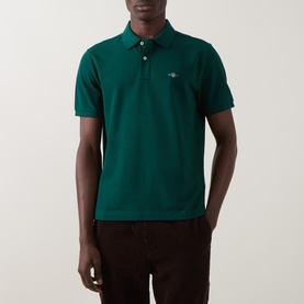 Model wearing Gant Regular Polo in dark emerald, short sleeves, piqu&eacute; cotton with logo badge, styled with dark brown trousers.