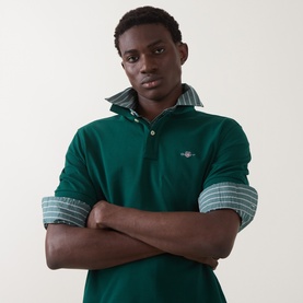 Model wearing Gant Regular Polo in dark emerald cotton, arms crossed, styled with a green checked shirt underneath.