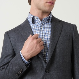 Model wearing Canali D8 Herringbone Single-Breasted Blazer in charcoal, styled with a blue check shirt. Notch lapels and button detail visible.