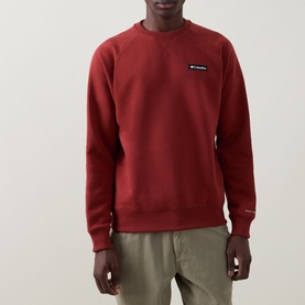 Model wearing Columbia crew neck sweatshirt in Red Oxide, with small chest logo badge, paired with khaki trousers.