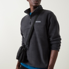 Model wearing Columbia black quarter-zip fleece pullover with small chest logo patch, styled with blue top and black crossbody bag.