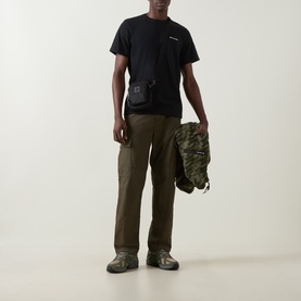 Model wearing Columbia CSC Logo Back Graphic T-Shirt in black with olive cargo trousers, carrying a camo jacket and black crossbody bag.