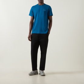 Model wearing Columbia CSC Logo Back Graphic T-Shirt in Phoenix Blue, round neck, short sleeves, styled with black trousers and grey trainers.