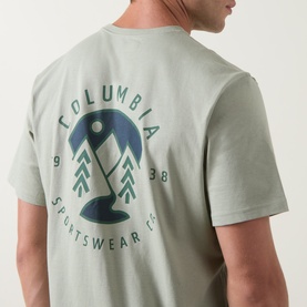 Model wearing Columbia CSC Outdoor Back Graphic T-Shirt in Safari green, showing circular mountain and tree logo graphic on back.