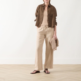 Model wearing Marc O Polo Linen Straight-Leg Trousers in Sandpaper with brown overshirt, vest top and dark thong sandals, holding woven clutch