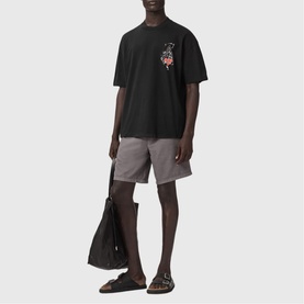 Model wearing All Saints Washed Black oversized T-shirt with chest graphic, styled with grey shorts and brown sandals, carrying a black tote bag.
