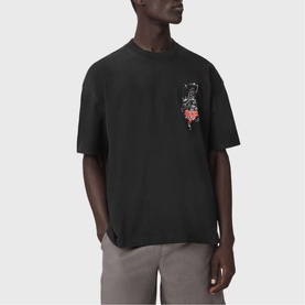 Model wearing All Saints washed black oversized T-shirt with small floral graphic print on chest, paired with grey trousers.