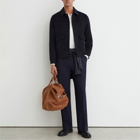 Model carrying a tan suede bag with gold hardware, styled with a navy jacket, white polo, and navy trousers