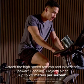 Model using a stationary exercise bike with a Dyson fan attachment, leaning forward in athletic wear in a dark wood-panelled studio.