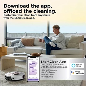 Shark robot vacuum on floor beside sofa, with man using SharkClean app on phone. Text highlights app-based home mapping and scheduling.