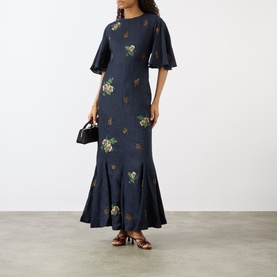 Model wearing Agua Bendita Ave Anhelo Dress in multicolour floral embroidery on navy, maxi length with flared hem, styled with strappy heels and mini