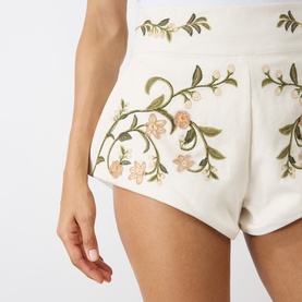 Model wearing Agua Bendita multicolour linen shorts with pink and green floral embroidery on white fabric, close-up detail view.